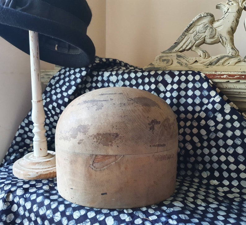 May include: A wooden hat block with a worn, natural finish. The block is round and has a slight indentation at the top. It is sitting on a blue and white patterned fabric.