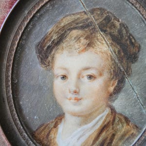 May include: Oval portrait miniature of a young person with light skin, brown hair, and a brown jacket. The portrait is set in a decorative, dark metal frame. A crack runs diagonally across the portrait.
