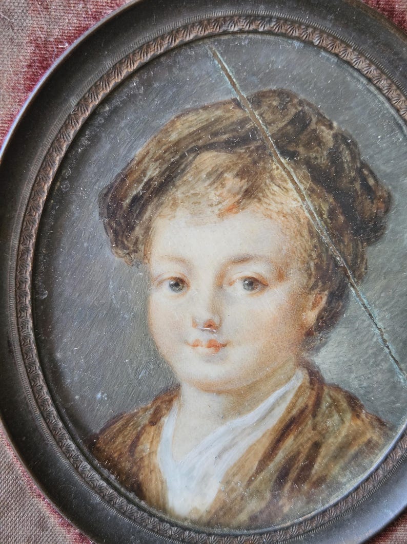 May include: Oval portrait miniature of a young person with light skin, brown hair, and a brown jacket. The portrait is set in a decorative, dark metal frame. A crack runs diagonally across the portrait.
