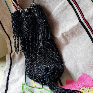 Può includere: Una borsetta vintage nera con perline, manico a catena e frange. La borsetta è fatta di piccole perline lucenti e pende da una catena. Lo sfondo è un tessuto dai toni neutri con dettagli floreali ricamati.