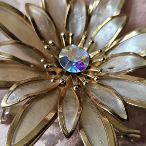 May include: A vintage flower brooch with gold-tone metal petals and a central iridescent crystal. The petals have a white enamel filling, and the brooch has a pin closure. The design is reminiscent of a daisy or sunflower.