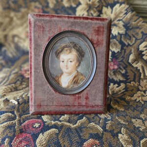 May include: A small, antique portrait of a child in a dark frame. The oval portrait features a young person with light skin, wearing a brown jacket and a dark hat. The frame is a rectangular shape with a red velvet texture.