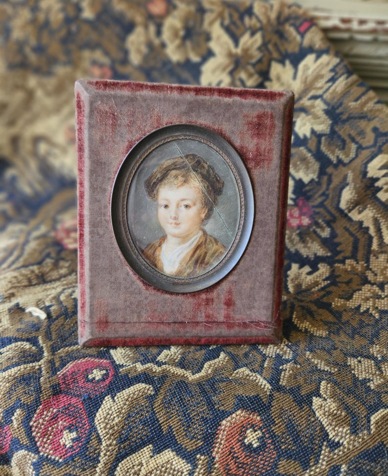 May include: A small, antique portrait of a child in a dark frame. The oval portrait features a young person with light skin, wearing a brown jacket and a dark hat. The frame is a rectangular shape with a red velvet texture.