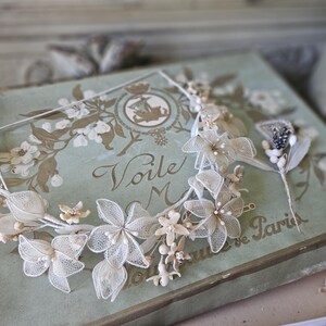May include: A delicate, floral headpiece with a matching hair accessory. The headpiece features white, lace-like flowers with pearl accents. The items rest on a vintage, light green box with floral and script details. The box reads "Voile M" and "Souvenir de Paris".