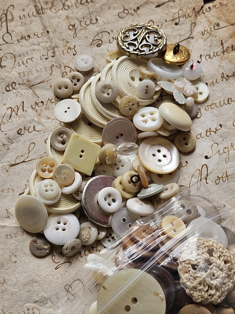 May include: A collection of vintage buttons in various shapes, sizes, and colors, including white, cream, and brown. Some buttons have a pearl-like finish. The buttons are arranged on a piece of paper with handwritten text.