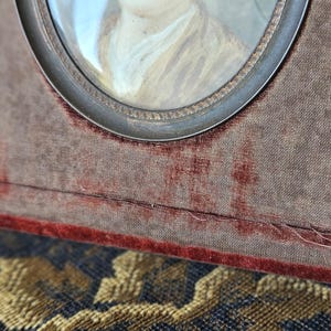 May include: Antique portrait miniature in a velvet-covered frame. The frame is a rectangular shape with a silver-toned metal oval frame around the portrait. The velvet is a muted brown color with a red border.