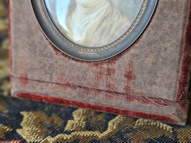 May include: Antique portrait miniature in a velvet-covered frame. The frame is a rectangular shape with a silver-toned metal oval frame around the portrait. The velvet is a muted brown color with a red border.