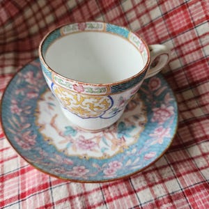 Op de afbeelding: Een vintage theekopje en schotelset met een bloemenpatroon. Het kopje en de schotel zijn wit met blauwe, roze en gele bloemenpatronen.