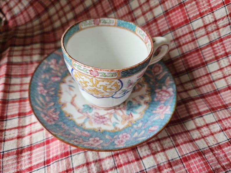 Op de afbeelding: Een vintage theekopje en schotelset met een bloemenpatroon. Het kopje en de schotel zijn wit met blauwe, roze en gele bloemenpatronen.