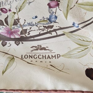 Puede incluir: Bufanda color crema con un diseño floral y botánico. Presenta acentos morados, azules y verdes, con el logotipo de Longchamp Paris. La bufanda tiene un borde rosa y escritura a mano.