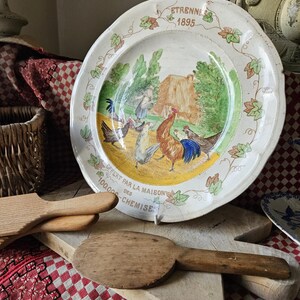 May include: A white ceramic plate with a yellow, green, and brown painted scene of roosters and hens. The plate has a decorative border of green leaves and the text "ETRENNES 1895" and "CHEZ PAR LA MAISON DES CHEMISE 100,000".
