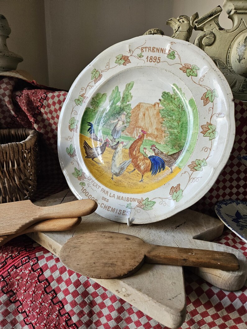 May include: A white ceramic plate with a yellow, green, and brown painted scene of roosters and hens. The plate has a decorative border of green leaves and the text "ETRENNES 1895" and "CHEZ PAR LA MAISON DES CHEMISE 100,000".