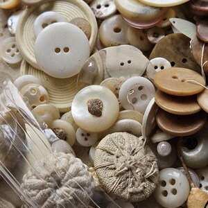 May include: A collection of vintage buttons in various shapes, sizes, and colors, including white, cream, and brown. The buttons are made of different materials, such as plastic, metal, and fabric. Some buttons have a decorative design, while others are plain.