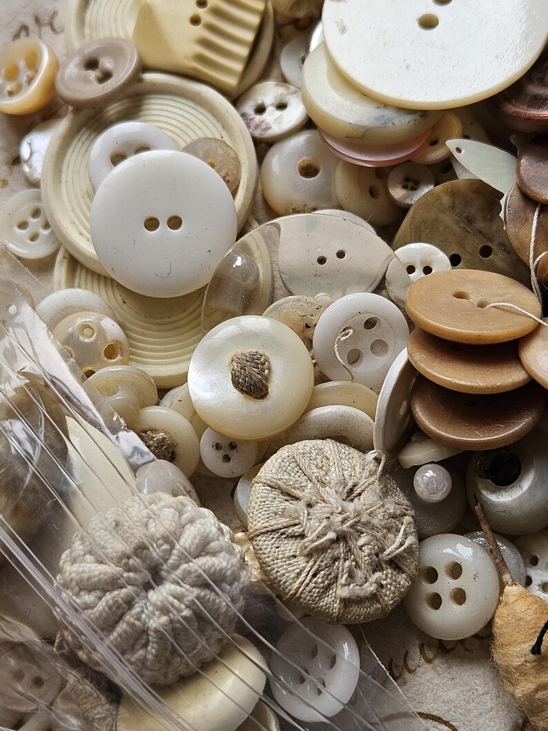May include: A collection of vintage buttons in various shapes, sizes, and colors, including white, cream, and brown. The buttons are made of different materials, such as plastic, metal, and fabric. Some buttons have a decorative design, while others are plain.