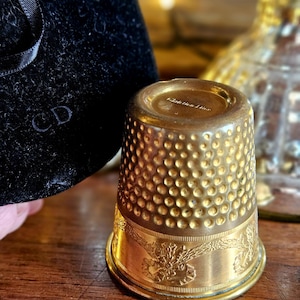 Könnte beinhalten: Ein goldfarbener Fingerhut mit strukturierter Oberfläche und eingraviertem floralem Design. Der Fingerhut trägt den Text "Christian Dior" auf der Oberseite. Ein schwarzer Samtbeutel mit "CD" ist teilweise im Hintergrund zu sehen.