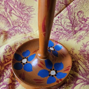 May include: A wooden stand with a round base painted with blue flowers and red accents. The stand has a tall, cylindrical wooden post.