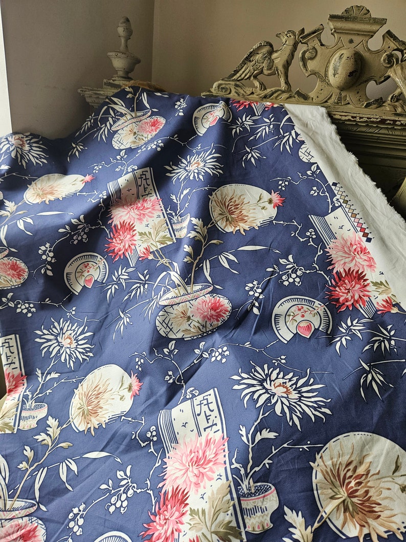 May include: A navy blue fabric with a floral pattern featuring pink chrysanthemums, white fans, and stylized vases. The design includes bamboo stalks and script-like elements, creating an Asian-inspired aesthetic. The fabric is draped, showcasing its texture and design.