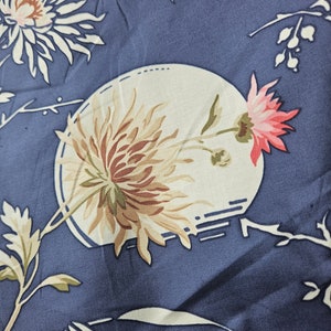 May include: Close-up of a blue fabric with a floral pattern. The design features large, stylized flowers in shades of cream, brown, and pink, set against a backdrop of blue. Delicate white branches and leaves complete the botanical theme.