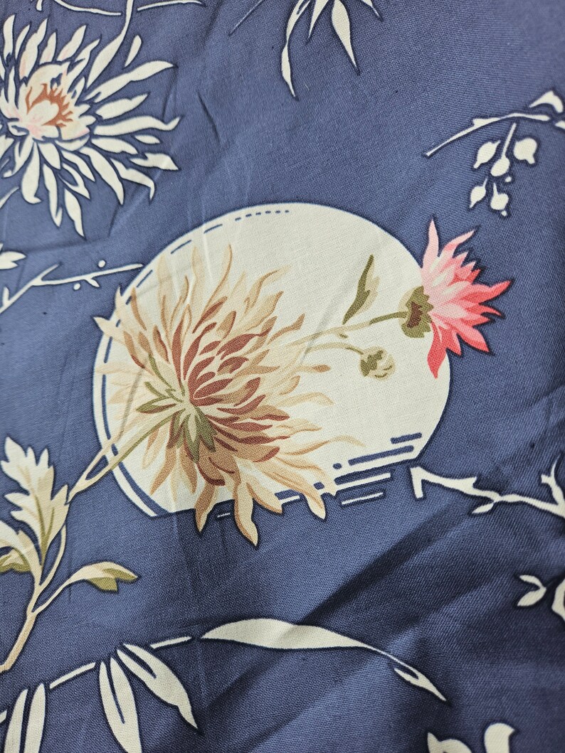 May include: Close-up of a blue fabric with a floral pattern. The design features large, stylized flowers in shades of cream, brown, and pink, set against a backdrop of blue. Delicate white branches and leaves complete the botanical theme.