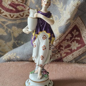 May include: A porcelain figurine of a woman in a purple and white dress, holding a white pitcher. The figurine has a gold-trimmed base and is decorated with floral details. The woman's dress has red floral accents.
