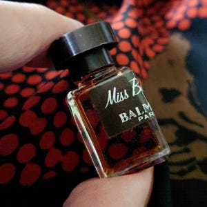May include: A small, rectangular perfume bottle with a dark brown cap. The bottle is filled with a dark amber liquid and has the words "Miss B BALM PAR" in white lettering. The bottle is held between fingers.