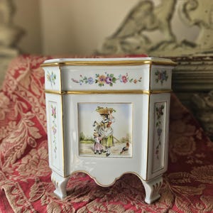Może przedstawiać: Biała porcelanowa donica ze złotymi obramowaniami i kwiatowymi akcentami. Donica przedstawia ręcznie malowaną scenę kobiety i dziecka. Donica ma sześciokątny kształt i spoczywa na małych nóżkach.