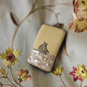Puede incluir: Un estuche compacto rectangular vintage con una tapa de color crema con un diseño de velero. El borde inferior está decorado con piezas iridiscentes en forma de mosaico. El estuche descansa sobre una tela bordada floral.
