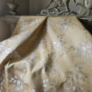 May include: A golden-yellow fabric with a floral pattern in silver and white. The fabric is draped over a surface, with the folds and textures visible. The floral design includes stylized leaves and blossoms.