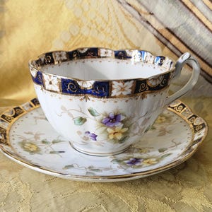 May include: A white teacup and saucer set with a floral design. The teacup has a blue and gold rim and a delicate handle. The saucer has a matching design. The set is on a gold-colored surface.