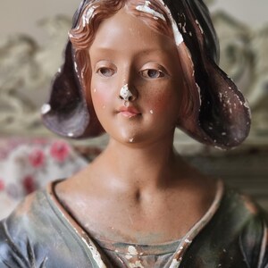 May include: A vintage statue of a young person with a brown complexion, wearing a dark hood and a dark green dress. The statue has visible signs of age, with chipped paint revealing the underlying material. The figure's face has a serene expression.