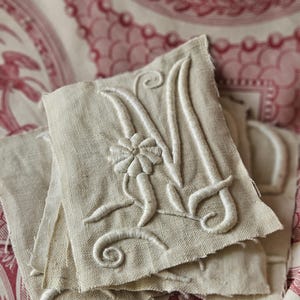May include: Close-up of several beige linen fabric squares with embroidered white capital letter "M" and floral details. The squares are stacked on top of each other, with a red and white patterned fabric in the background.