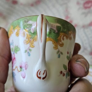 Op de afbeelding: Een porseleinen theekop met een witte basis en een groene rand, versierd met gele krullen en roze bloemmotieven. Het handvat is wit met een dunne oranje lijn in het midden en een oranje decoratief motief aan de onderkant.