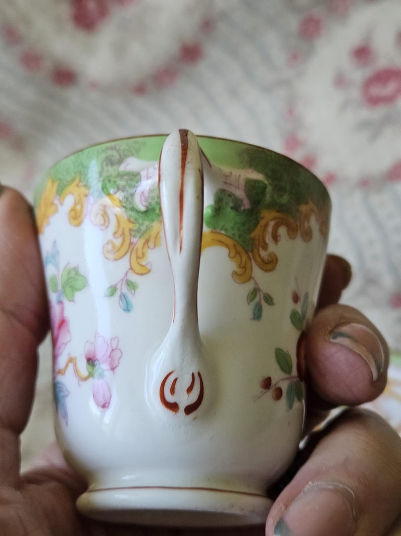 Op de afbeelding: Een porseleinen theekop met een witte basis en een groene rand, versierd met gele krullen en roze bloemmotieven. Het handvat is wit met een dunne oranje lijn in het midden en een oranje decoratief motief aan de onderkant.