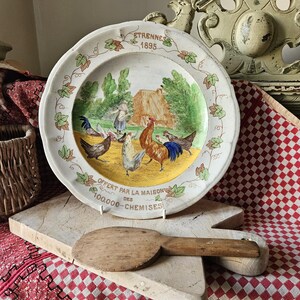 May include: A white ceramic plate with a colorful painted scene of chickens and a rooster. The plate has a scalloped edge and a yellow center with the text "OFFERT PAR LA MAISON DES 100,000-CHEMISES" and "ETRENNES 1895".