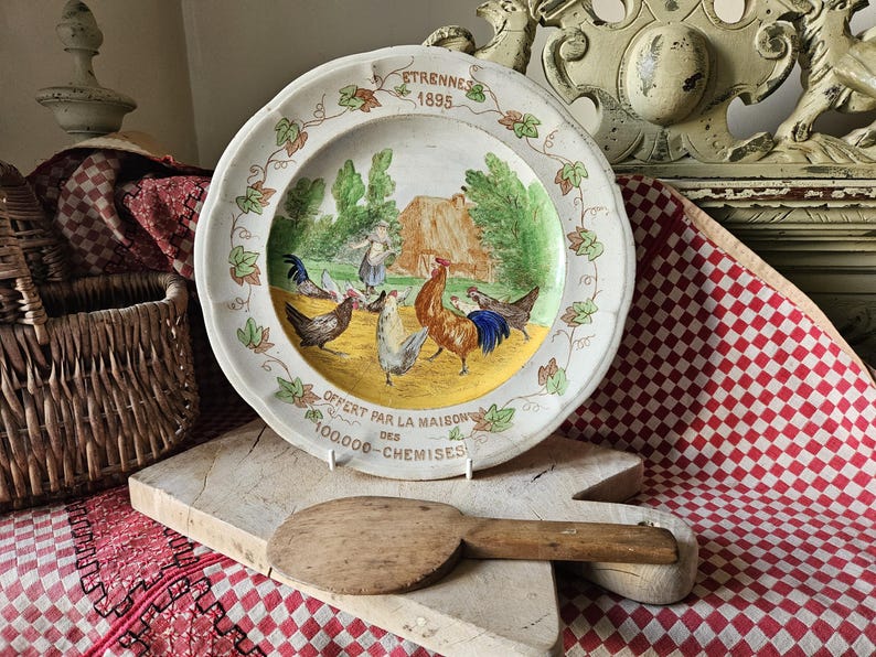May include: A white ceramic plate with a colorful painted scene of chickens and a rooster. The plate has a scalloped edge and a yellow center with the text "OFFERT PAR LA MAISON DES 100,000-CHEMISES" and "ETRENNES 1895".