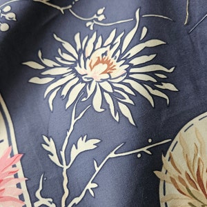 May include: Close-up of a fabric with a floral pattern. The design features a large white flower with a brown center and pink accents, set against a dark blue background. Other floral elements and decorative shapes are also visible.