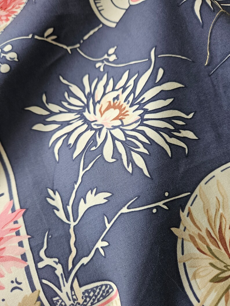 May include: Close-up of a fabric with a floral pattern. The design features a large white flower with a brown center and pink accents, set against a dark blue background. Other floral elements and decorative shapes are also visible.