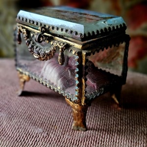 May include: An antique glass and metal jewelry box with a hinged lid. The box has a rectangular shape with ornate gold-colored metal trim and legs. The glass panels reveal a soft, purple interior. The lid is a pale blue color.