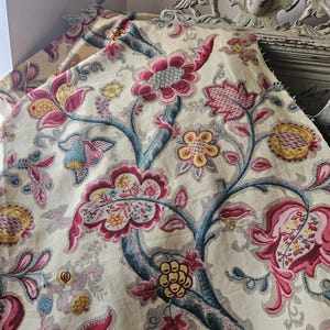 May include: A cream-colored fabric with a floral pattern. The flowers are pink, red, yellow, and blue. The fabric has a vintage look.