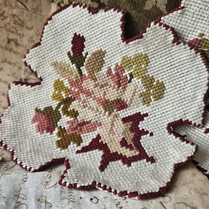 May include: A white fabric with a burgundy border, featuring a floral design with pink, white, and green flowers. The fabric is stitched with a cross-stitch pattern.