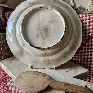 May include: A large, white ceramic plate with a raised rim and a slightly textured surface. The plate is decorated with a simple, faded design. The plate is sitting on a wooden surface.
