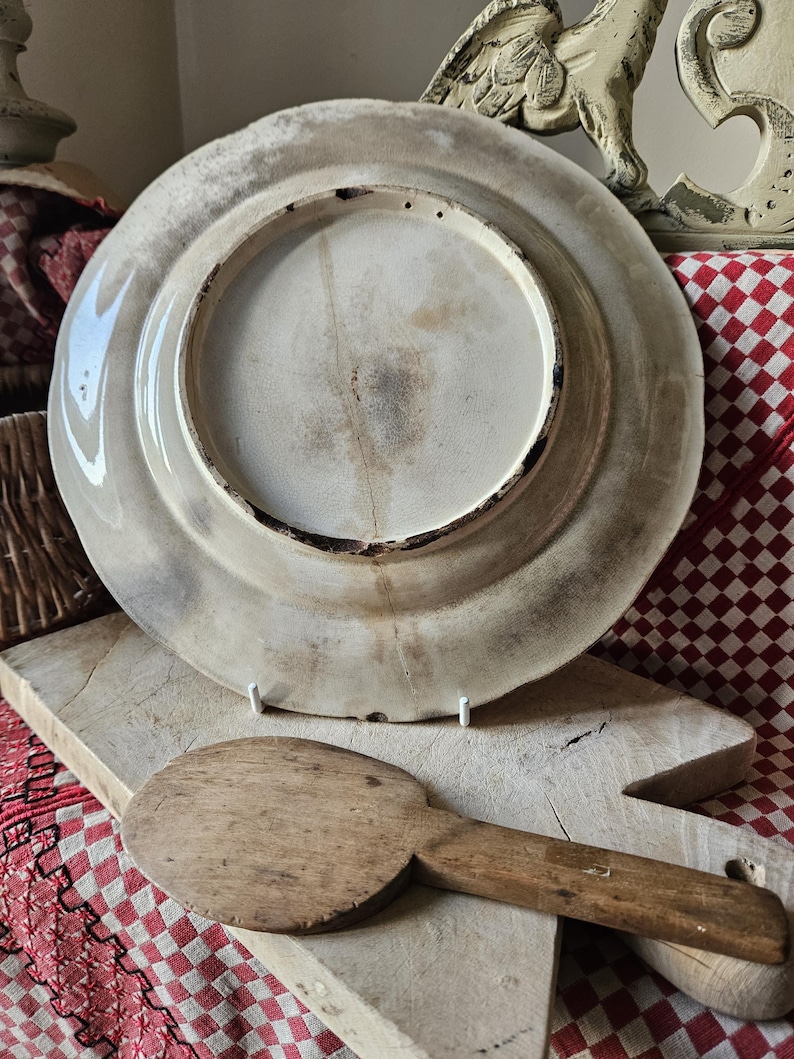 May include: A large, white ceramic plate with a raised rim and a slightly textured surface. The plate is decorated with a simple, faded design. The plate is sitting on a wooden surface.