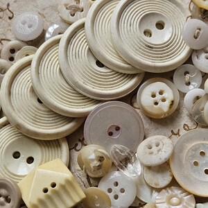 May include: A collection of vintage buttons in various shapes, sizes, and colors, including white, cream, and beige. Some buttons have a textured surface, while others are smooth. The buttons are arranged on a piece of paper with writing on it.