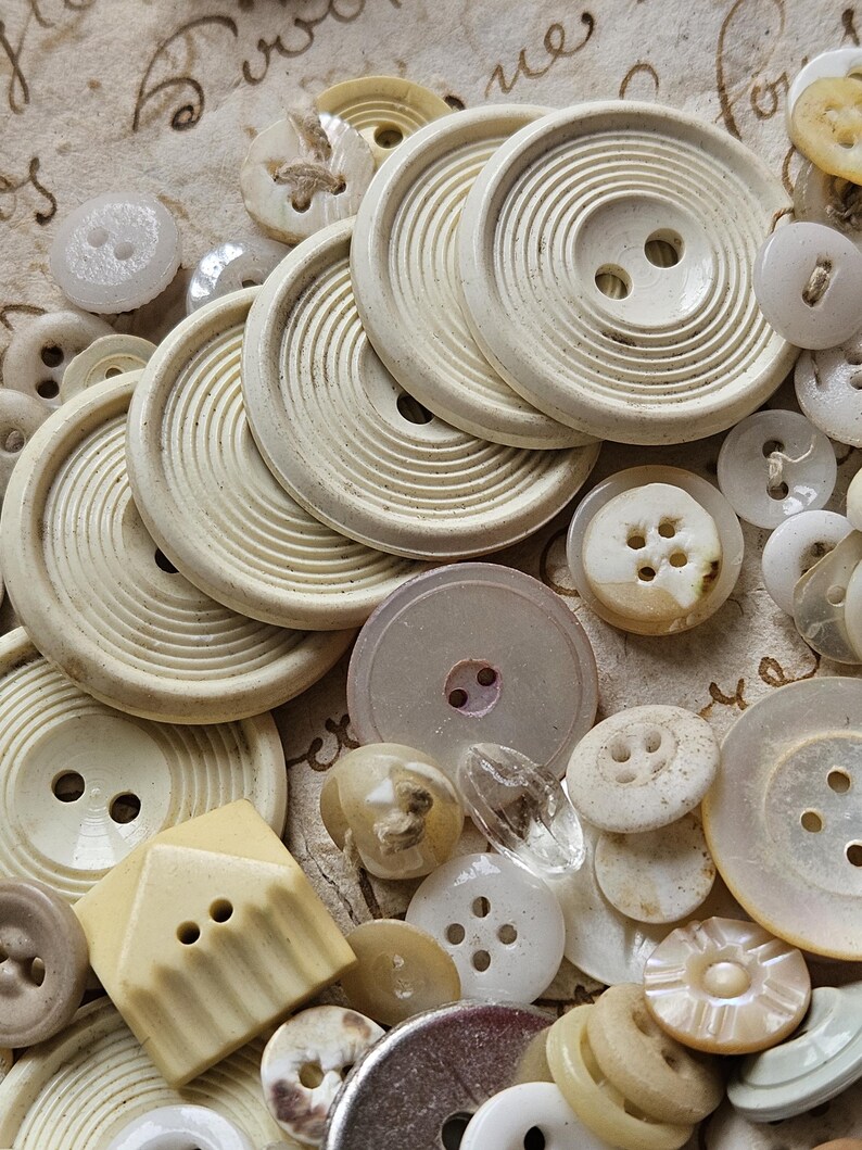 May include: A collection of vintage buttons in various shapes, sizes, and colors, including white, cream, and beige. Some buttons have a textured surface, while others are smooth. The buttons are arranged on a piece of paper with writing on it.