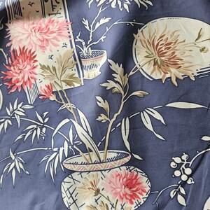 May include: Close-up of a fabric with a floral pattern in shades of pink, cream, and brown on a dark blue background. The design features stylized flowers, bamboo, and decorative pots, creating an Asian-inspired aesthetic. The fabric has a soft, flowing appearance.