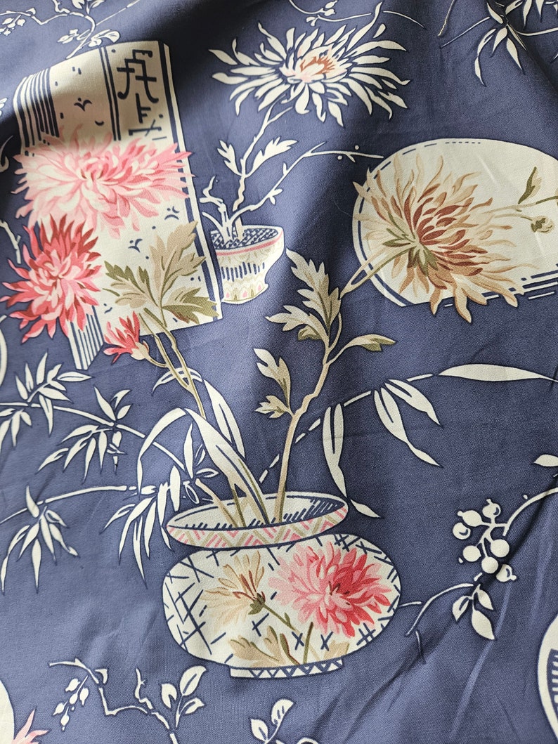 May include: Close-up of a fabric with a floral pattern in shades of pink, cream, and brown on a dark blue background. The design features stylized flowers, bamboo, and decorative pots, creating an Asian-inspired aesthetic. The fabric has a soft, flowing appearance.