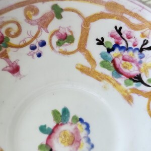 Op de afbeelding: Een witte porseleinen kom met een handgeschilderd bloemmotief in roze, blauwe en groene tinten. De binnenzijde is versierd met gouden ranken en een groene geometrische rand, afgewerkt met een goudkleurige boord.