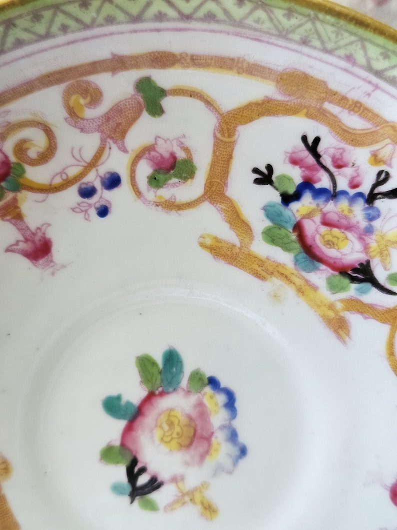 Op de afbeelding: Een witte porseleinen kom met een handgeschilderd bloemmotief in roze, blauwe en groene tinten. De binnenzijde is versierd met gouden ranken en een groene geometrische rand, afgewerkt met een goudkleurige boord.