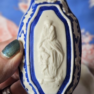 Puede incluir: Una figura religiosa de cerámica blanca y azul. La figura tiene un borde azul y un centro blanco con un relieve de una figura vestida con una túnica. Los bordes están decorados con un motivo floral. La figura es sostenida por una persona.