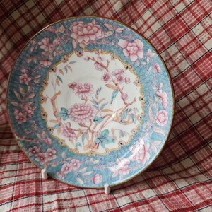 Op de afbeelding: Een vintage porseleinen schotel met een blauw en wit bloemenmotief. De schotel heeft roze bloemen en groene bladeren op een lichtblauwe achtergrond. De rand van de schotel is versierd met een gouden rand.